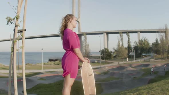 Beautiful Girl in Dress Standing in Skate Park with Longboard alt
