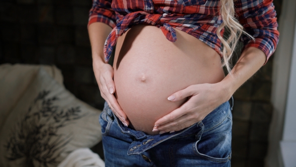 of Charming Belly and Hands of a Happy Young Pregnant Woman in Blue Jeans and Plaid Shirt alt