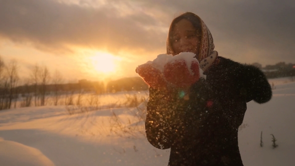 Winter Portrait of a Beautiful Young Woman Blowing Snowflakes and Smiling in  at Magic Sunset