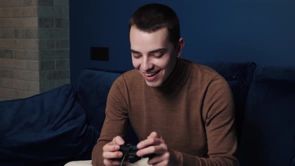 Happy Excited Young Caucasian Man Gamer Holding Controller Playing Video Games Sitting on Sofa alt