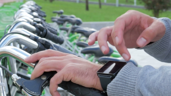 A Man Checks the Messages on the Smart Watch on a Bike Parking Lot. He's Going To Rent a Bike