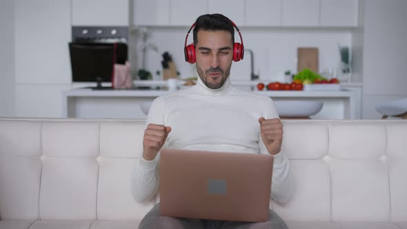 Satisfied Intelligent Middle Eastern Man in Headphones Making Victory Gesture Sitting on Couch with alt
