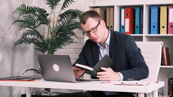 Office Worker Businessman or Entrepreneur in Glasses and a Suit Works with a Notebook and a Laptop alt