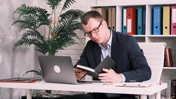 Office Worker Businessman or Entrepreneur in Glasses and a Suit Works with a Notebook and a Laptop