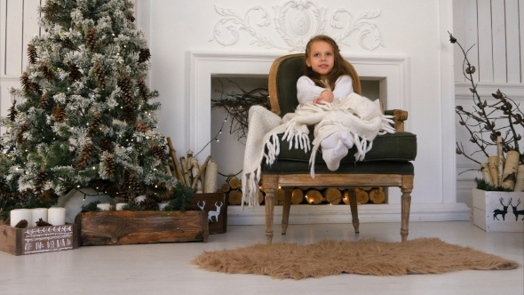 Smiling Girl Wrapped in a Blanket Sitting on a Cozy Chair and Waiting for Santa alt