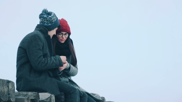 Loving Couple Sitting on a Wooden Bridge, on a Background of Gray Sky of Snow, and Talking.