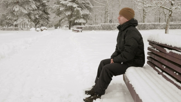 A Young Man Sits on a Bench in Winter Park and Admiring the Snow