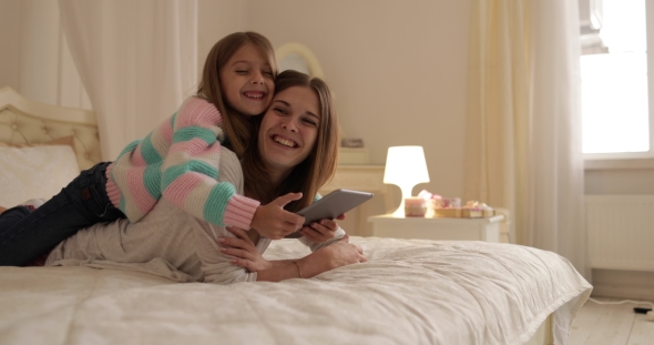 Mother With Daughter Lying On Bed Using Tablet Computer Happy Smiling Young Family alt