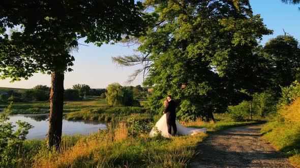 Newlyweds on Their Wedding Day are Standing By the Lake in a Green Park alt