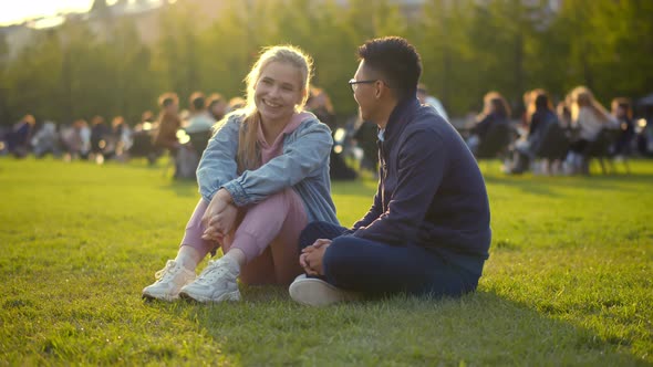 Young Multiethnic Couple Talking and Laughing Sitting on Grass in Park alt