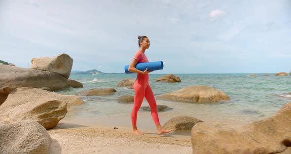 Woman Walking on the Beach for Morning Exercise She Carry Blue Yoga Mat and Wears Pink Sports Suit alt