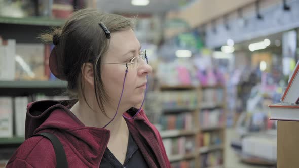 Serious Caucasian Woman with Glasses on Chain Buys a Book in Bookstore
