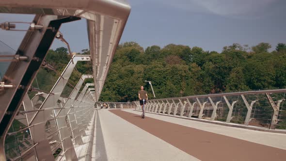Modern Young Man Rides an Electric Scooter Across the Pedestrian Bridge Overlooking the City alt
