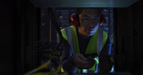 Asian male it technician wearing headphones and using tablet checking computer server alt