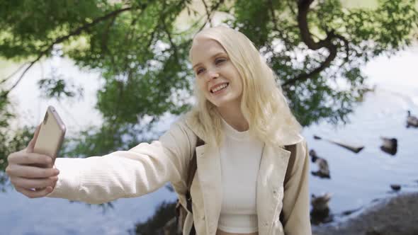 Cute Woman Tourist Happily Smiles and Takes a Selfie on the Background of the Lake with Ducks alt