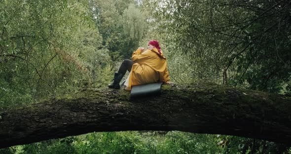 Hiker in Red Bandana and Yellow Raincoat Sits on Trunk of Fallen Tree in Forest