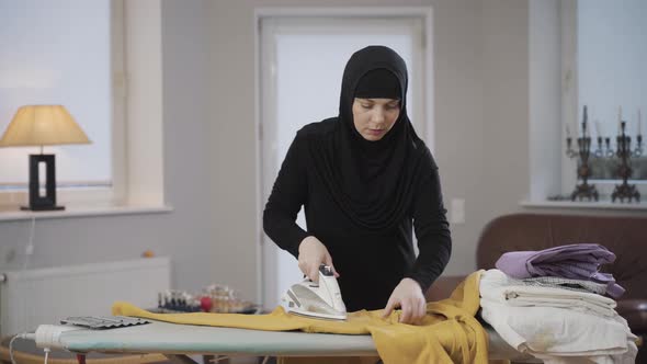 Portrait of Concentrated Muslim Woman in Hijab Ironing Yellow Pullover. Housewife Doing Housework alt