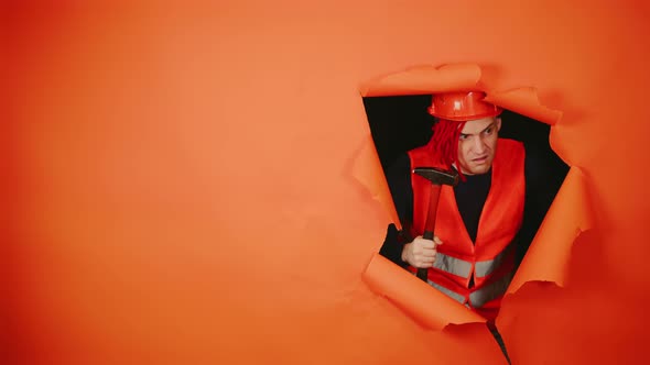 Shocked Male Construction Worker in Overalls with Hammer Looking Out of Hole of Orange Background alt