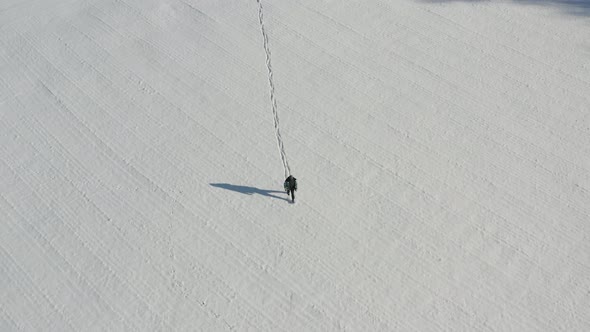 Birdseye aerial view of lonely man walking on snow capped field leaving trail on sunny winter day, h alt