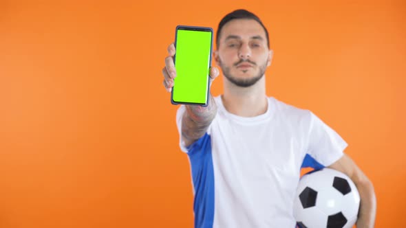 Football Fan In White Blue Shirt and Ball Show Green Screen Smartphone Orange Background  alt