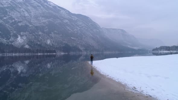 Man walks alone, snowy Bohinj lake reflecting mountains in distance, aerial flyover shot. Slovenia. alt