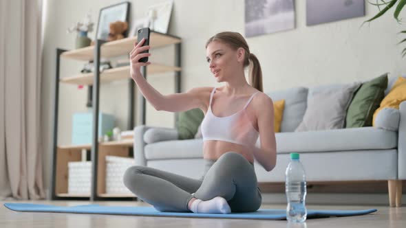 Young Woman Talking on Video Call on Smartphone While Sitting on Yoga Mat alt