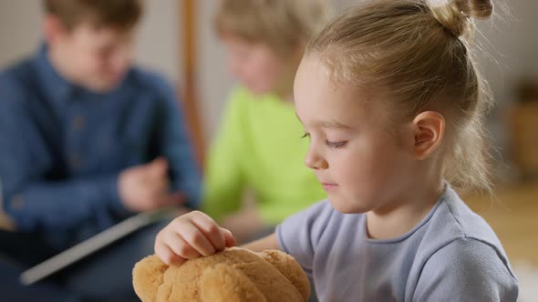 Closeup Side View of Little Pretty Girl Hugging Teddy Bear with Blurred Boys Talking at Background alt