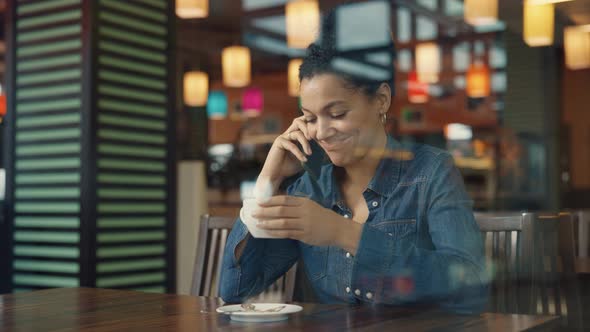 View Through Glass Showcase of Young African American Woman Talking on Phone and Enjoying Coffee in alt