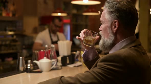 Close-up of an adult gray-haired man with a beard drinking whiskey in a bar