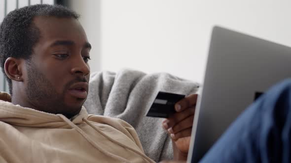 Young Man At Home Lying On Sofa With Laptop Using Credit Card To Shop Online