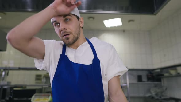 Front View Tired Man in Cook Uniform Rubbing Forehead Thinking As Colleague Passing at Background alt