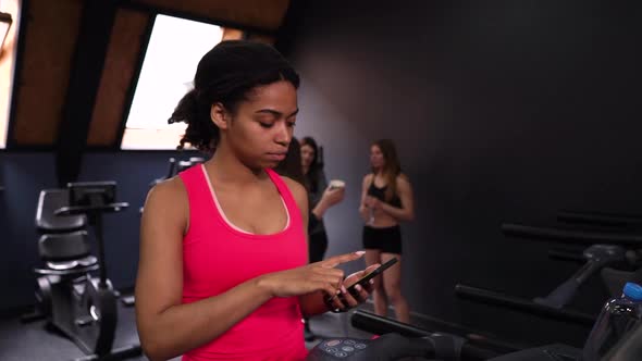 Black woman using smartphone on treadmill in gym alt