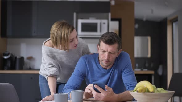 Husband Messaging Online on Smartphone Distracted By Wife Talking Sitting at Table in Kitchen alt