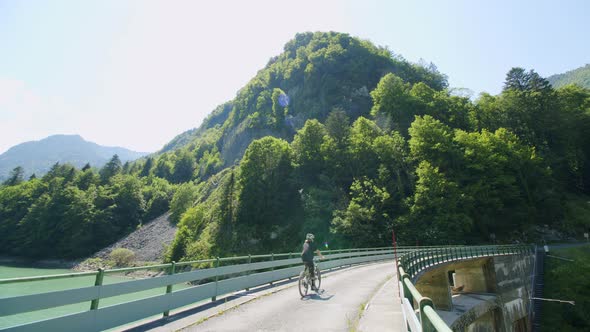 Mountain biker rides across a dam with a tall mountain in front of him alt