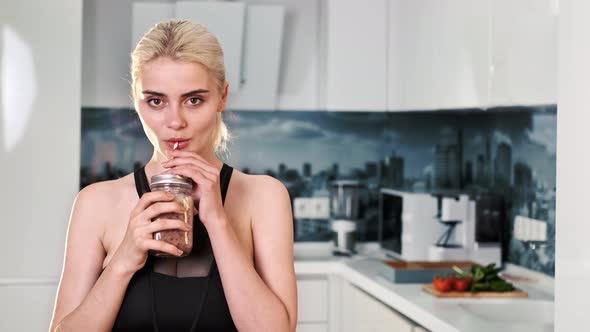 Young blonde smiling woman looking into the camera and drinking a healthy drink alt