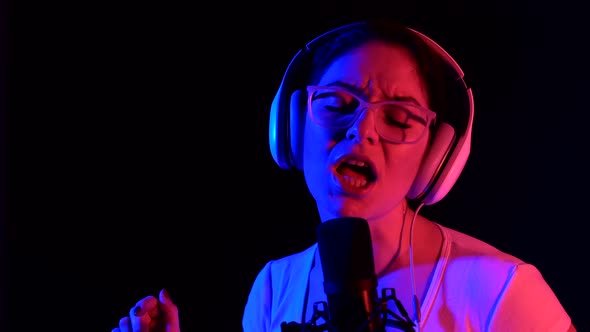 Caucasian Woman in Glasses and Headphones Sings Into a Microphone in Neon Light on a Black alt