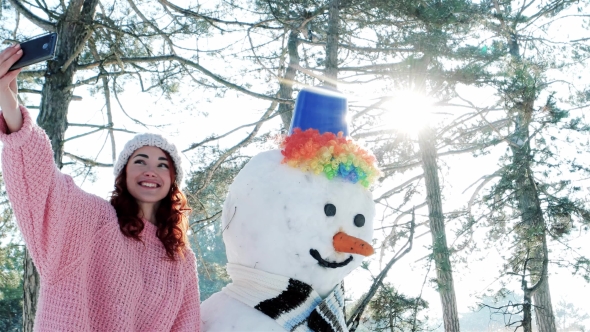 Winter Selfie, Cute Girl Making Photo with Snowman, a Mobile Phone in the Hand of Young Woman Making alt
