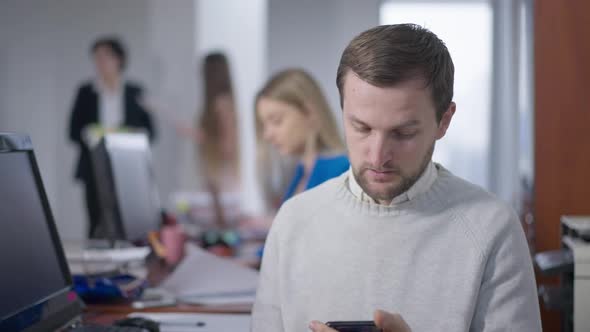 Brunette Caucasian Man Sneezing Surfing Internet on Smartphone in Office and Blurred People Leaving alt