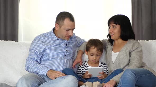 The Family Is Sitting on the Couch and Looking Intently at the Tablet Screen. Dad, Mom, and a Small alt