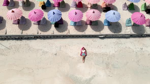 Aerial top down of girl lying and relaxing on tropical beach surrounded by colorful sunshades. alt