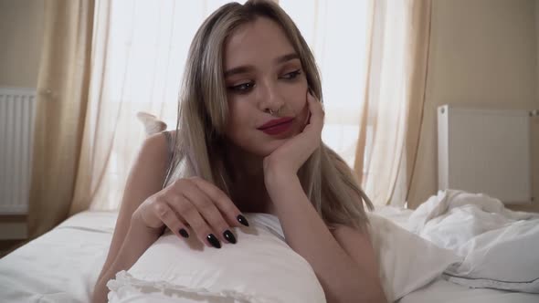 Pretty Girl with Bright Makeup and Pierced Nose Is Lying on the Bed with Her Legs Up alt