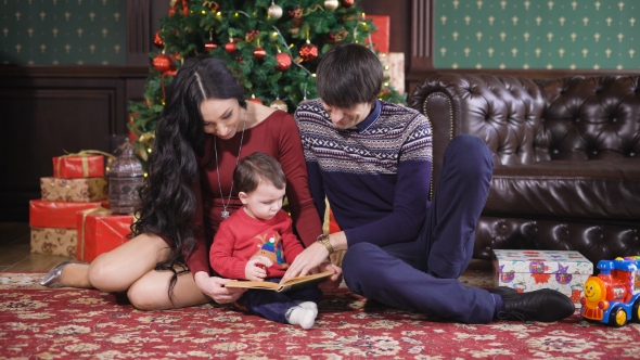 Christmas Room. The Family Has Gathered Around a New Year Tree. The Kid Holds the Book in Hand alt
