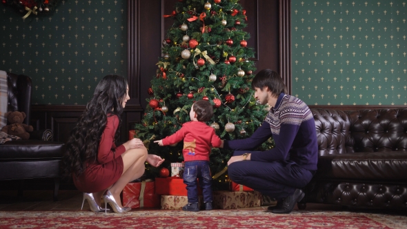 Young Family Sits Near the Rich Christmas Tree. It Is Decorated Garland, Toys, Ribbons, Bows. Mom alt