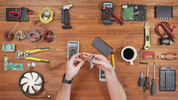 Man Repairing a Mobile Phone. Checks Parts Inside the Device. Wooden Table Top View.