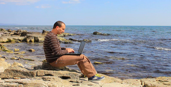 Man Working With Laptop On A Rocky Beach