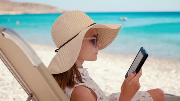 Lady in Hat Reads Electronic Book on EReader on Beach at Seaside in Summer
