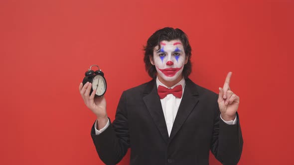 Portrait of a Strange Clown in a Suit with an Alarm Clock in His Hands on an Isolated Red Background