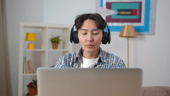 Young Asian Man in Headphones Watching Education Webinar on Laptop at Home alt