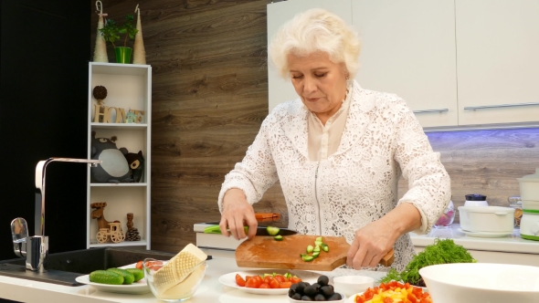 Old Gray-haired Woman Cut Cucumbers in the Kitchen.