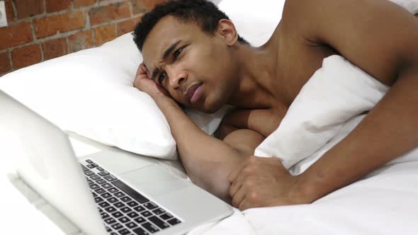 Pensive African Man in Bed Working on Project on Laptop alt